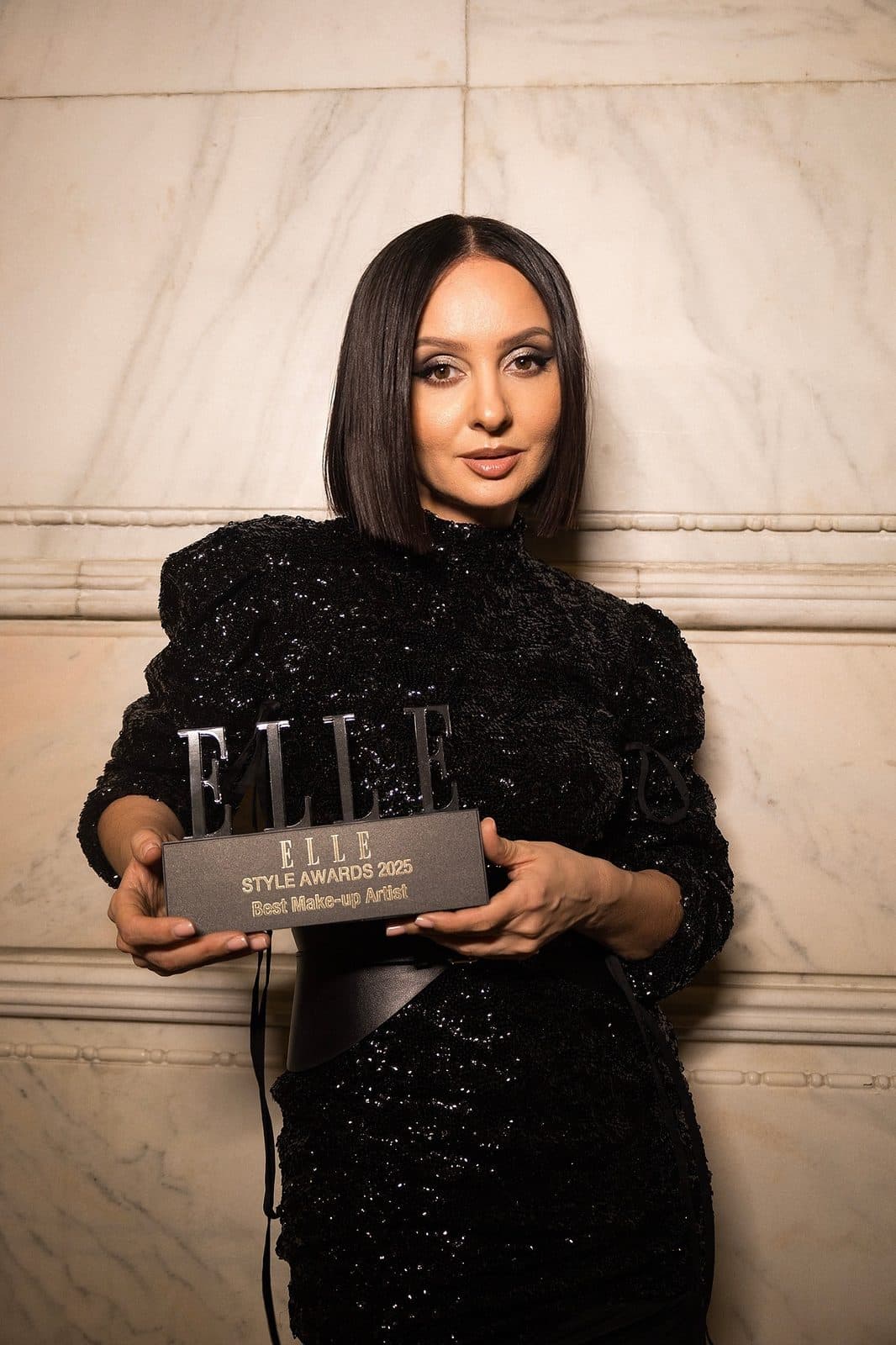 Tania Cozma ține trofeul ELLE Style Awards 2025 pentru cel mai bun make-up artist, în fața unui perete de marmură.