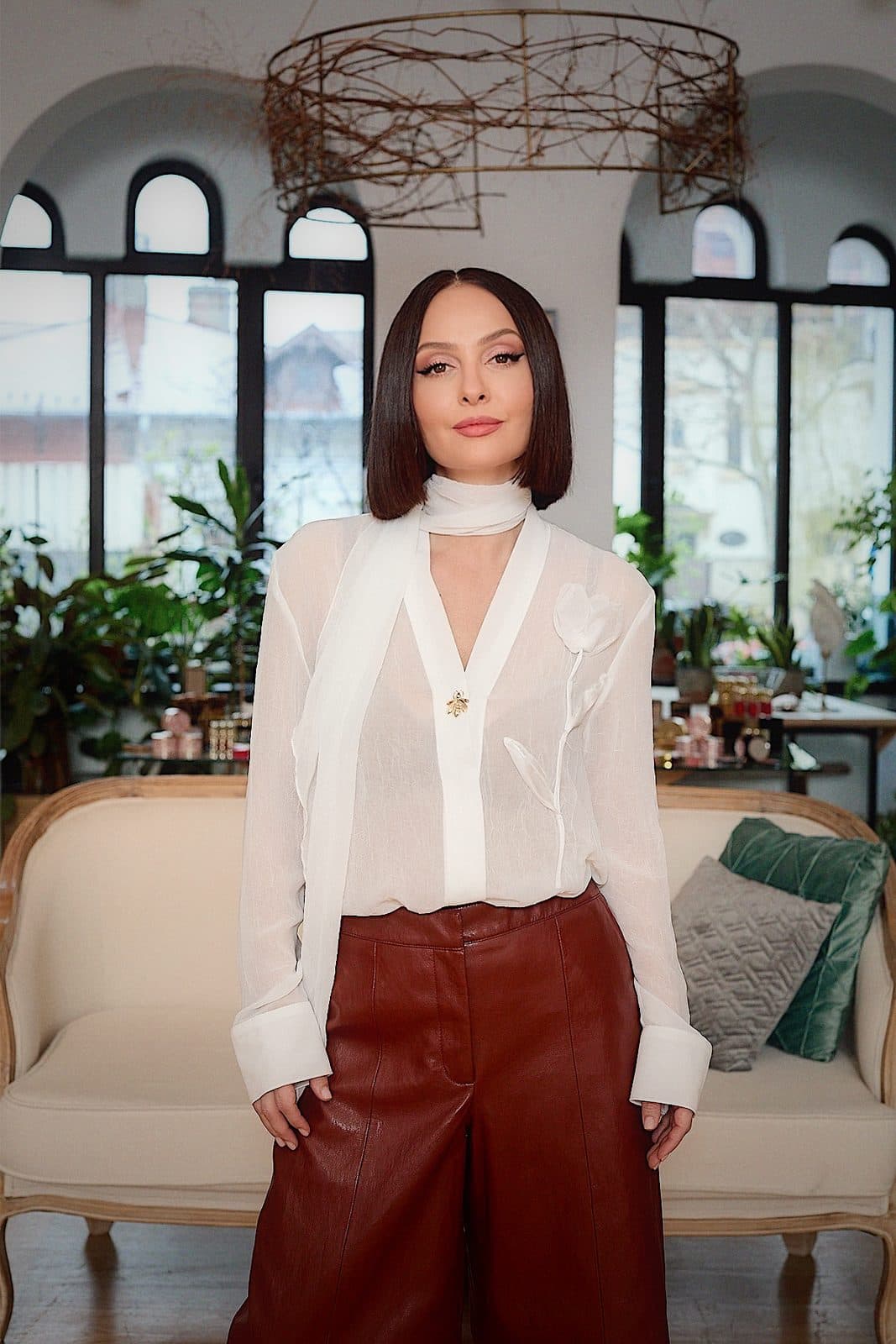 Tania Cozma în bluză albă transparentă și pantaloni din piele vișinii, în picioare într-un loft cu ferestre arcuite.