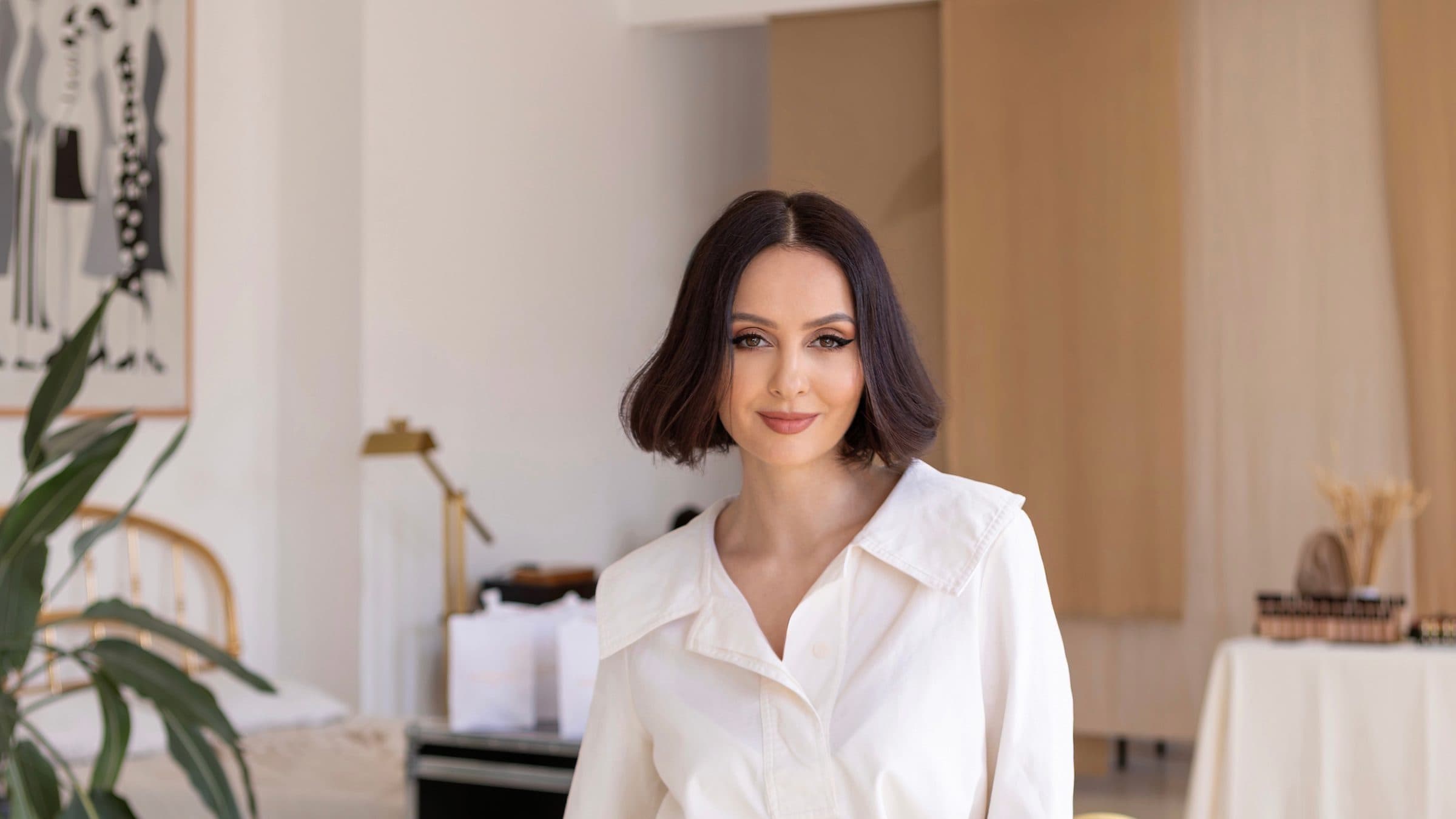 Tania Cozma, în cămașă albă, zâmbește sprijinită de o masă cu fonduri de ten și produse de machiaj, într-un studio luminos.