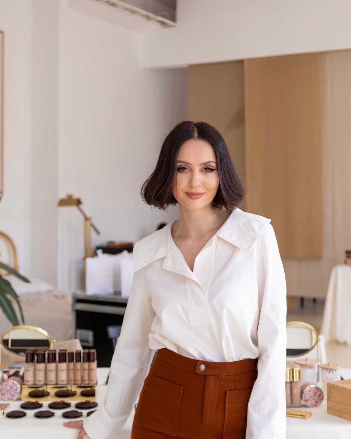 Tania Cozma, în cămașă albă, zâmbește sprijinită de o masă cu fonduri de ten și produse de machiaj, într-un studio luminos.
