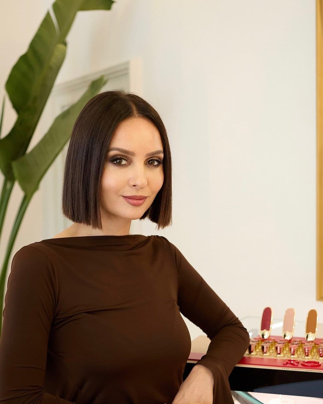 Portret al Taniei Cozma în rochie maro, sprijinită de o vitrină cu rujuri, într-un salon crem.