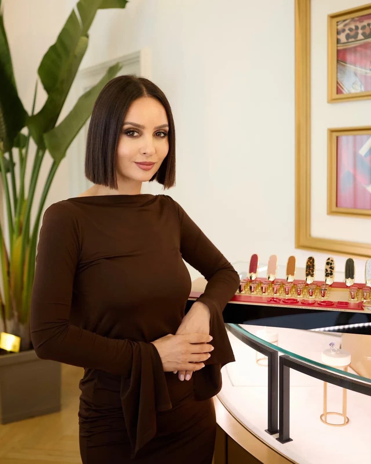 Tania Cozma, în rochie maro cu mâneci evazate, sprijinită de o vitrină cu rujuri, într-un salon luminos cu o plantă mare.